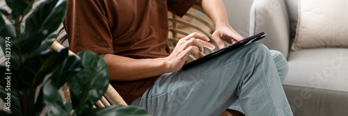 Technology Concept The male with his casual T-shirt and jeans sitting comfortably on the wooden chair and doing touchscreen for checking the web browser on the iPad