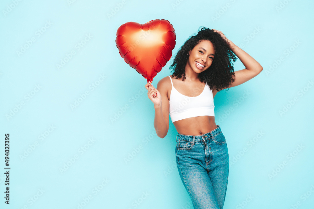 Beautiful black woman with afro curls hairstyle. Smiling model dressed in white summer dress. Sexy carefree female posing near blue wall in studio. Tanned and cheerful. Holding heart air balloon