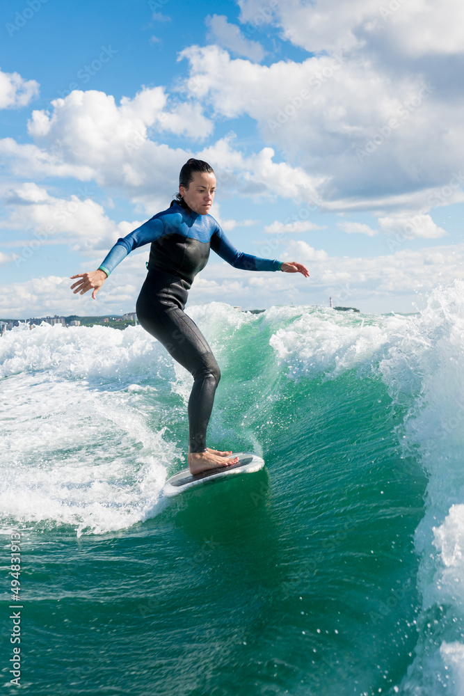 Young athletic female in wetsuit riding on endless waves behind a boat on sunny day. Woman practice wakeboarding, carving behind the baot. Watersport concept.