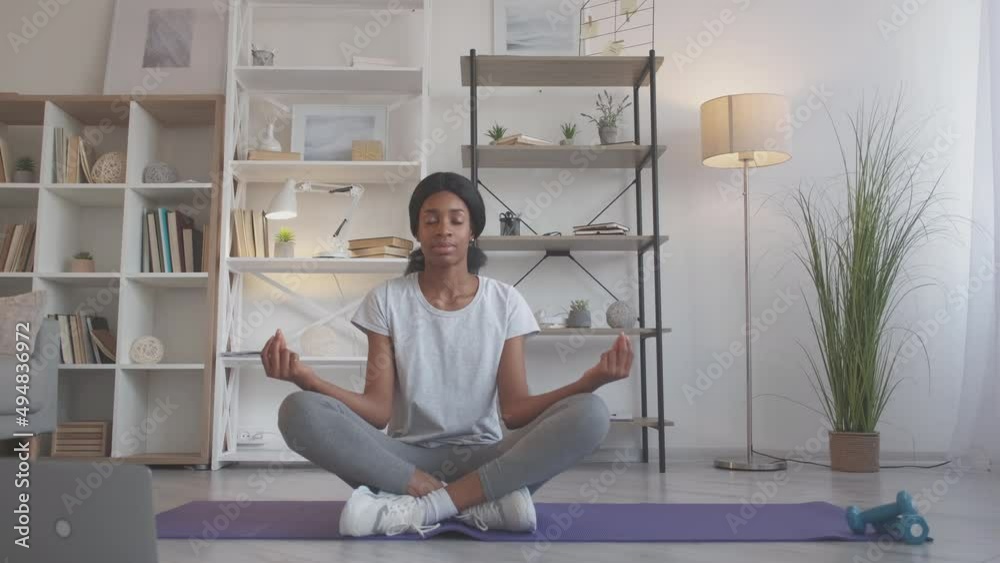 Home meditation. Indoors yoga. Stress relief. Peaceful mindful woman in sportswear relaxing on fitness mat in lotus pose at living room. Home meditation. Indoors yoga. Stress relief. Peaceful mindful woman in sportswear relaxing on fitness mat in lotus pose at living room.