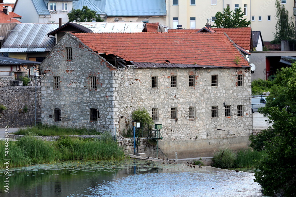 Large abandoned old building with traditional stone walls and concrete frame windows without glass covered with broken destroyed red roof tiles and metal sheets next to calm river and uncut grass
