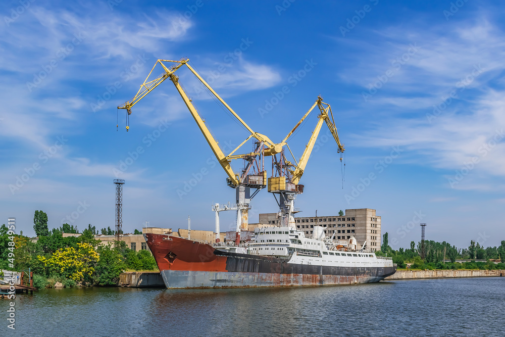 Mykolaiv, Ukraine - July 26, 2020: A large ship against the background of two port cranes on the Inhul River in Mykolaiv. View from the pedestrian bridge to the old rusty abandoned vessel