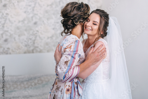Bride closes her eyes hugging mother tender. Mom congratulates the bride with a marriage and hugs. Happy bride with mother on background wall in home. Bride morning preparation. photo