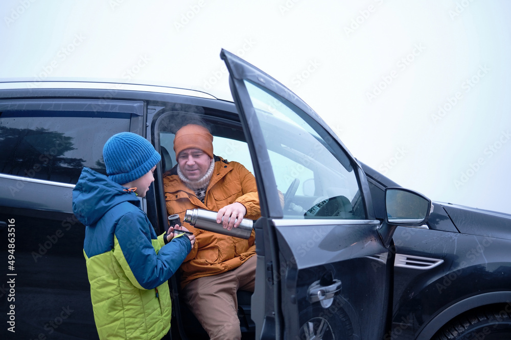 Naklejka premium Happy father and son Sitting together in a new car on a journey. Family are resting on the side of the road on a road trip. Child drinks hot tea from thermos. Happy family travels.