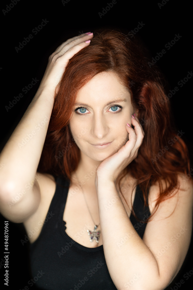 close-up of an attractive girl with red hair and blue eyes wearing a black tank top holding her head in her hands against a black background