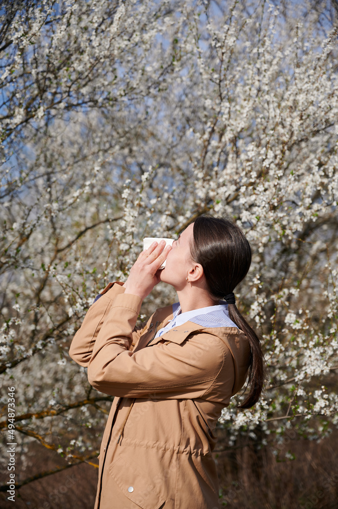 Woman allergic suffering from seasonal allergy at spring, posing in blossoming garden at springtime.Young woman sneezing and blowing nose in front of blooming tree. Spring allergy concept Woman allergic suffering from seasonal allergy at spring, posing in blossoming garden at springtime.Young woman sneezing and blowing nose in front of blooming tree. Spring allergy concept