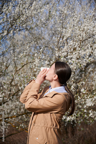 Woman allergic suffering from seasonal allergy at spring, posing in blossoming garden at springtime.Young woman sneezing and blowing nose in front of blooming tree. Spring allergy concept