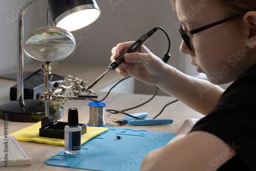 A teenager in glasses learns to solder with a soldering iron, holds a soldering iron in his hands and sits at the table