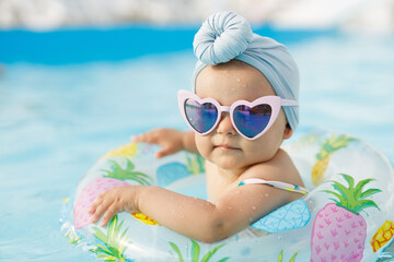 Cute funny toddler girl in colorful swimsuit and sunglasses relaxing on inflatable toy ring floating in pool have fun during summer vacation in tropical resort. Child having fun in swimming pool. 