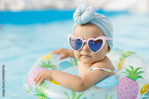 Cute funny toddler girl in colorful swimsuit and sunglasses relaxing on inflatable toy ring floating in pool have fun during summer vacation in tropical resort. Child having fun in swimming pool. 