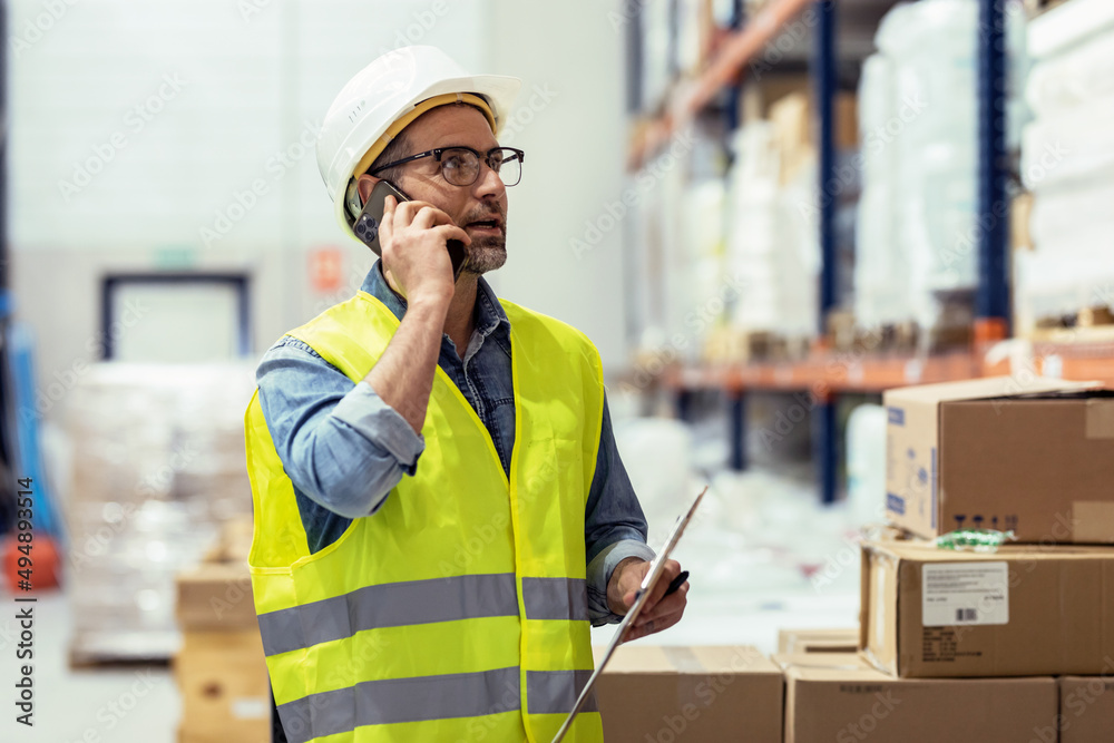 Mature worker talking with smartphone while passing list of material that has arrived at the store warehouse. Mature worker talking with smartphone while passing list of material that has arrived at the store warehouse.