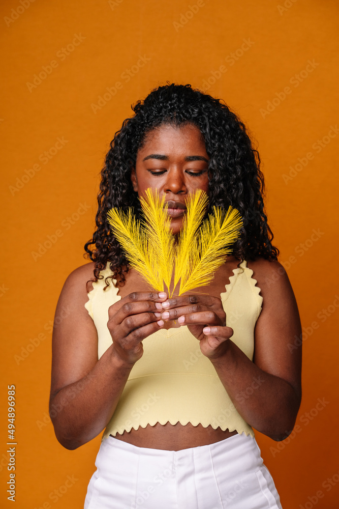 Mid waist portrait of afro woman blowing yellow soft feathers. Vertical front view of native american woman holding tribal feathers in orange background. People and ethnic culture.
