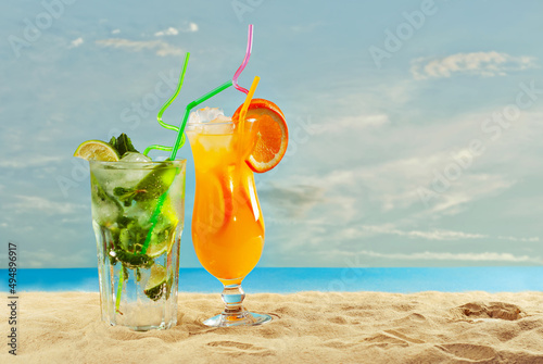 Orange and green cocktails on the beach. Alcoholic drink with ice, orange, mint and lime on the sand. Sunset, sea and sky in the background. Copy space and free space for text near the glass.