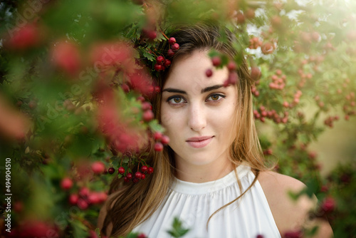 A beautiful young girl with long brown hair and brown eyes against a background of a green tree with bright red berries. Autumn portrait of a girl with professional makeup