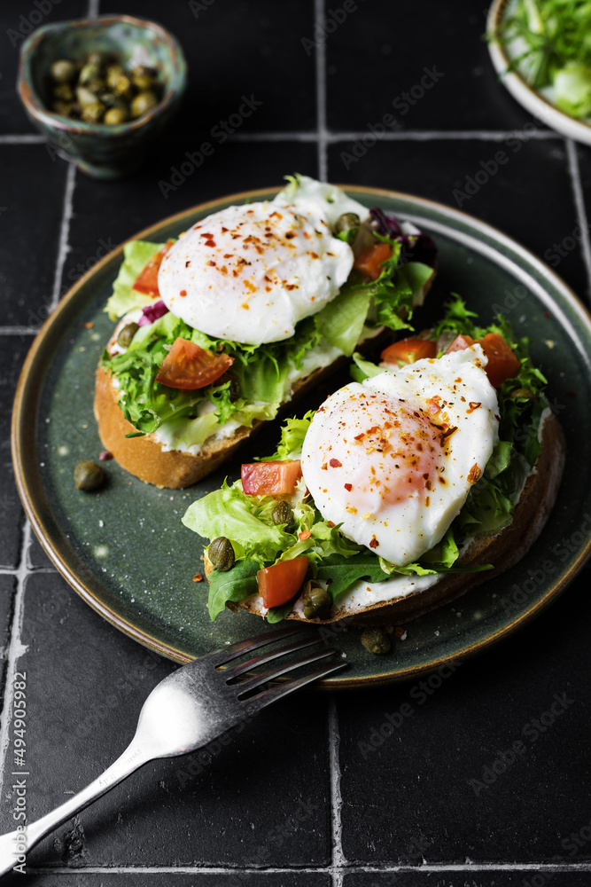 Toast with poached egg, lettuce, tomato and capers on a dark background, close up Toast with poached egg, lettuce, tomato and capers on a dark background, close up