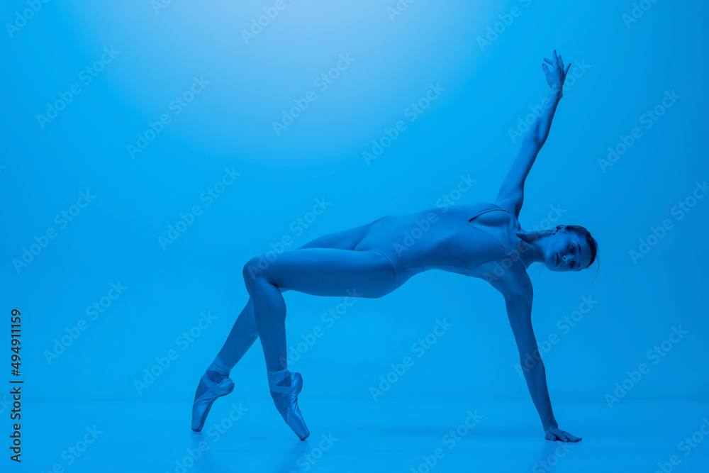 Portrait of beautiful graceful ballet dancer posing isolated on blue studio background in neon light. Art, motion, action, flexibility concept.