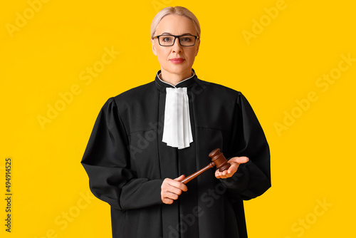 Mature female judge in robe with gavel on yellow background