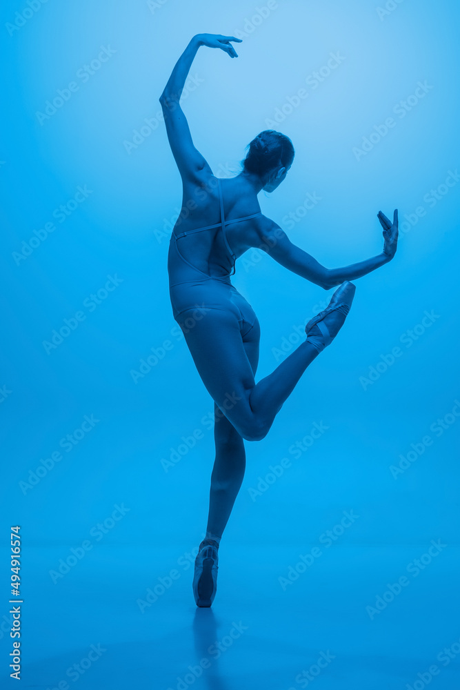 Back view of young and graceful ballet dancer isolated on blue studio background in neon light. Art, motion, action, flexibility, inspiration concept.