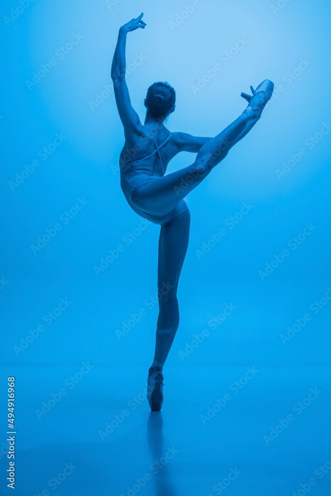 Back view of young and graceful ballet dancer isolated on blue studio background in neon light. Art, motion, action, flexibility, inspiration concept.