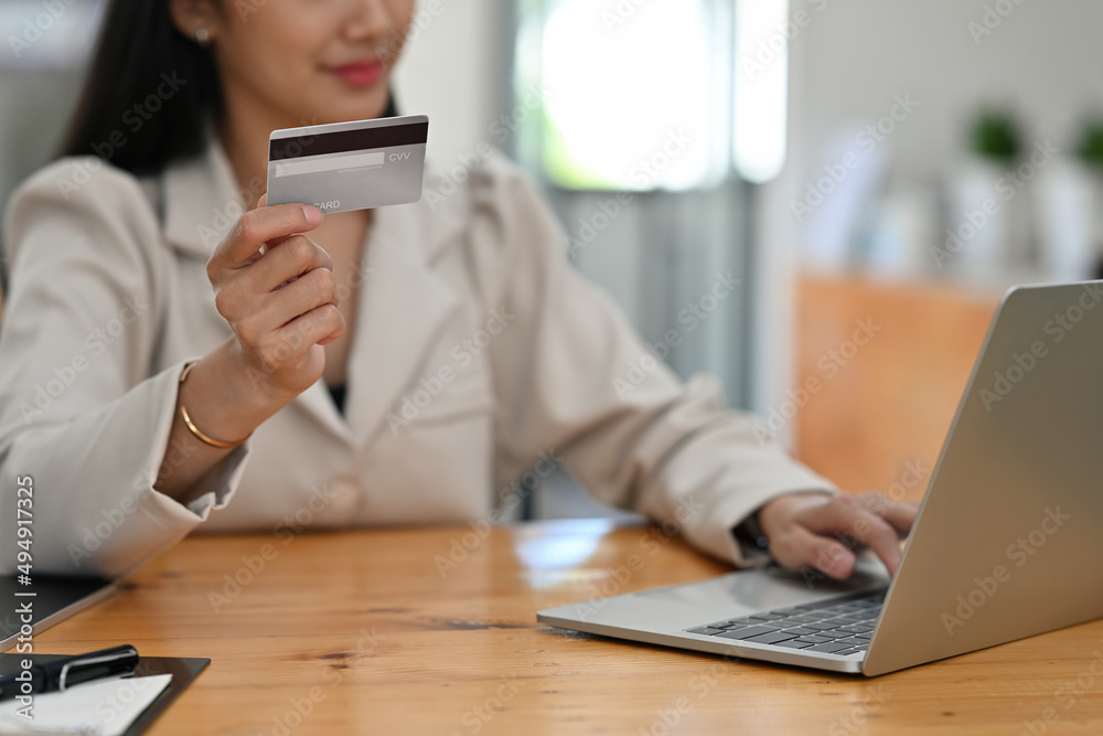 Cropped view of a smiling Asian woman sitting in the office holding a credit card and typing on a laptop, for finance and technology concept.