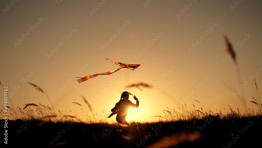 Happy child. Girl with flying kite in park. Child runs at sunset on grass in park. Silhouette of girl with flying kite. Child launches flying kite into sky in the wind. Silhouette of child in meadow