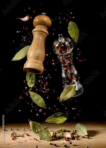 Fototapeta Naklejka Na Ścianę i Meble -  Two spice mills with peppercorns and laurel bay leaves on black flying over table.