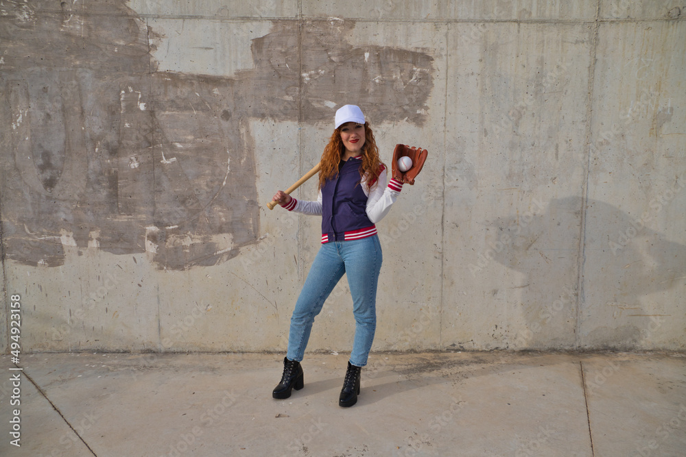 young and beautiful red-haired woman with baseball cap, jacket and glove with baseball bat resting on her shoulder on grey cement background. Sport and recreation concept.