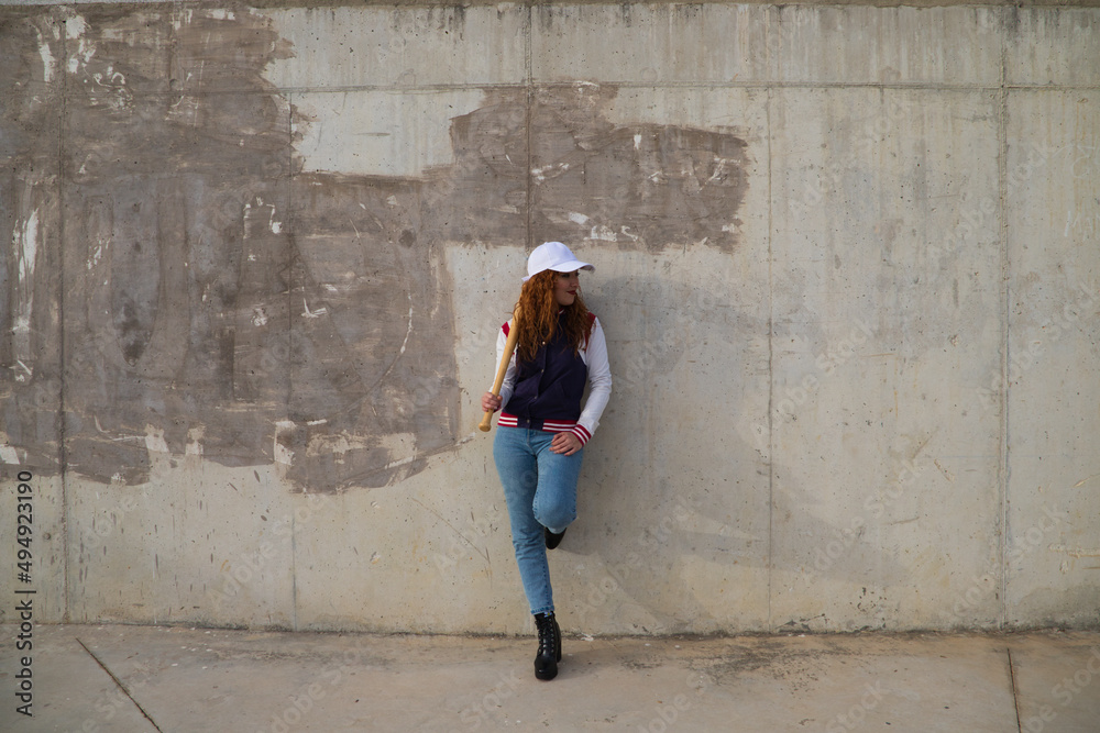 young and beautiful red-haired woman with baseball cap, jacket and glove with baseball bat resting on her shoulder on grey cement background. Sport and recreation concept. young and beautiful red-haired woman with baseball cap, jacket and glove with baseball bat resting on her shoulder on grey cement background. Sport and recreation concept.