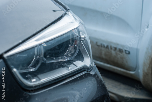 Headlight of an SUV close-up on the background of a white pickup. Dirty SUV sergo (metallic) colors
