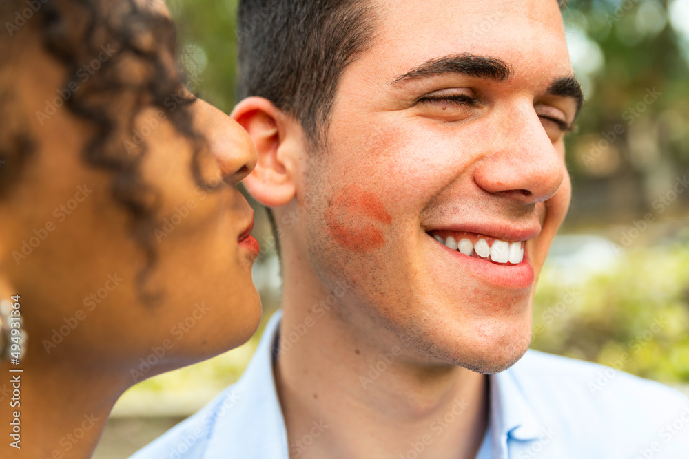 Multiethnic Young people kiss on cheek with lipstick – portrait of two people kissing on cheek – woman kissing her boyfriend -  happy loving woman kissing boy friend on cheek  - obrazy, fototapety, plakaty 