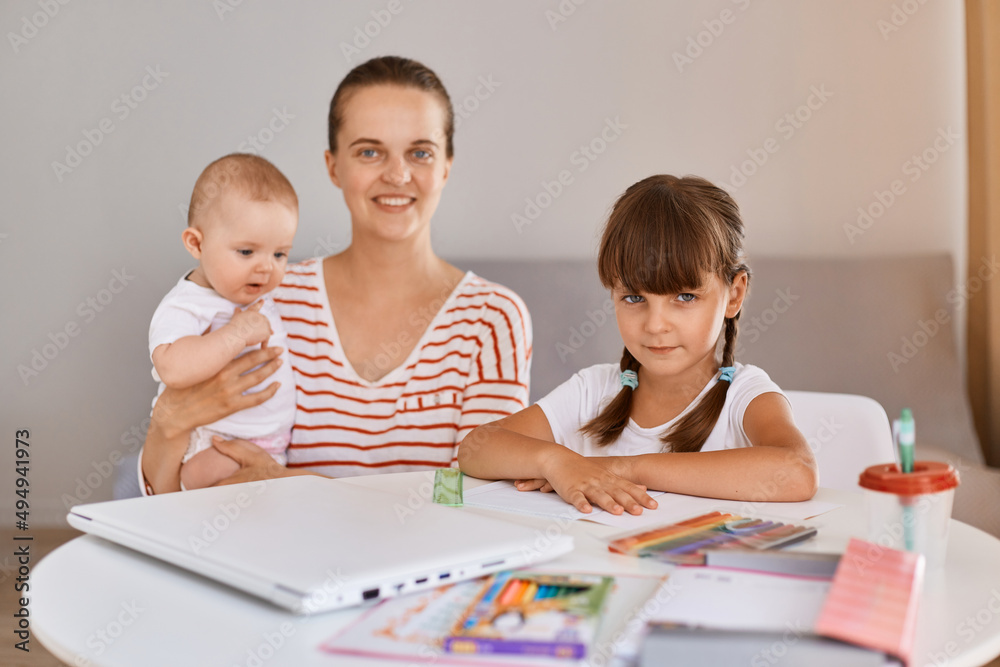 Indoor shot of positive optimistic young mother sitting with schoolgirl and infant daughters at table, doing school tasks at home, looking at camera. Indoor shot of positive optimistic young mother sitting with schoolgirl and infant daughters at table, doing school tasks at home, looking at camera.