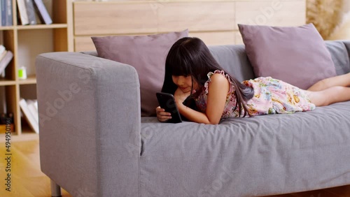 Cute little girl uses smartphone while prone at the sofa in the living room. Child surfing the internet on computer, browses through internet and watches cartoons online. photo