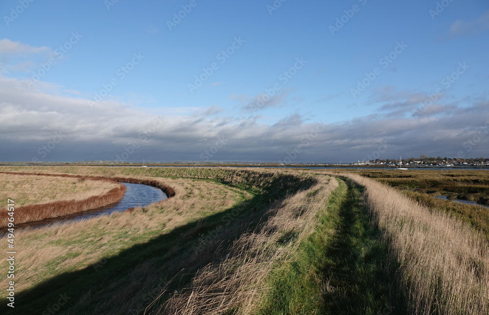 A beautiful scenic view along a curved grassy footpath through Old Hall Marshes nature reserve in Essex, UK with Mersea Island on the horizon. 