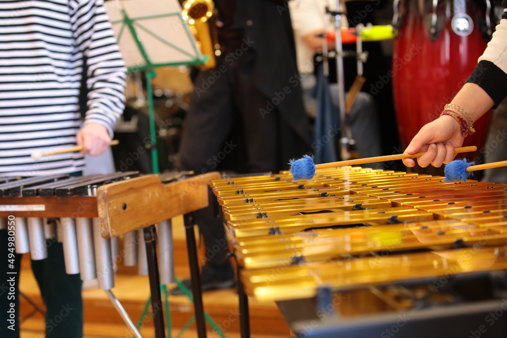 A person with hammers in his hands plays percussion in a youth jazz band with a player on saxophone drums piano rehearsing a concert standing on the school stage A person with hammers in his hands plays percussion in a youth jazz band with a player on saxophone drums piano rehearsing a concert standing on the school stage