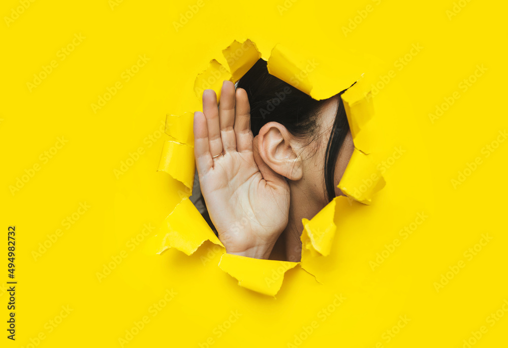 Close-up of a woman's ear and hand through a torn hole in the paper. Bright yellow background, copy space. The concept of eavesdropping, espionage, gossip and tabloids. - obrazy, fototapety, plakaty 