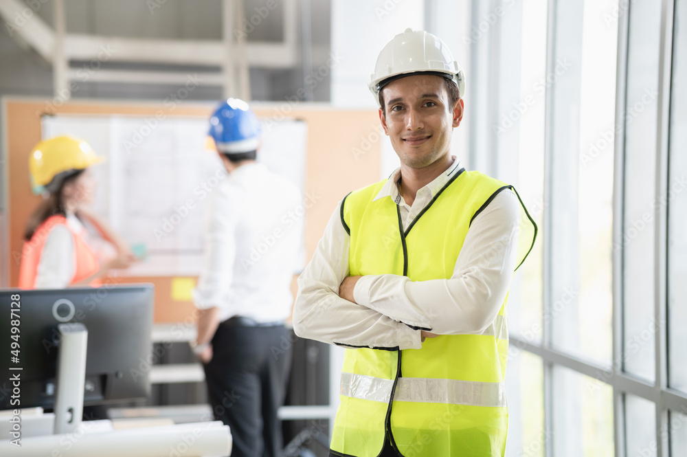 Portrait photo of young smart Hispanic engineer wearing safety helmet standing indoor of company office or construction site with colleague discussing about work in distance. Portrait photo of young smart Hispanic engineer wearing safety helmet standing indoor of company office or construction site with colleague discussing about work in distance.