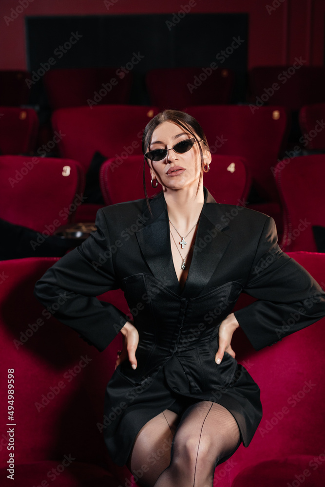 Fototapeta premium A model wearing all black sitting on a red cinema chair with nice sunglasses looking at camera