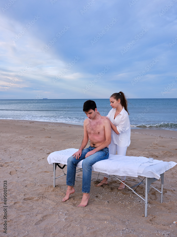 Full length view of a young man with bare torso sitting on a massage table receiving chiromassages on the beach in Valencia by a young chiromassage therapist.