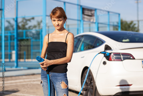 Beautiful young girl next to an electric car. Posing and holding a charging cable.