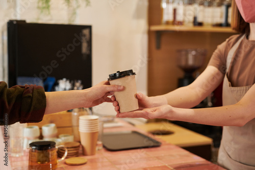 Medium section side view shot of unrecognizable female barista wearing mask standing at counter giving cup of coffee to customer