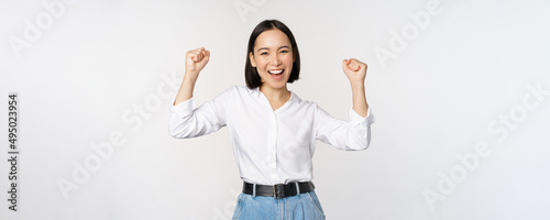 Enthusiastic asian woman rejoicing, say yes, looking happy and celebrating victory, champion dance, fist pump gesture, standing over white background