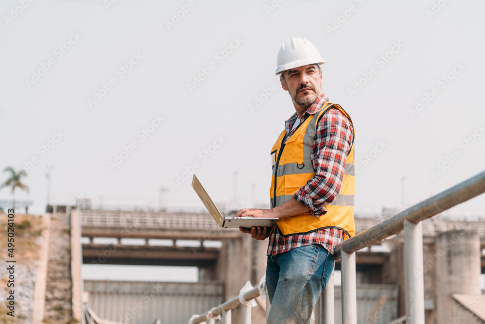 Portrait of power engineer wearing safety jacket and hardhat with laptop computer working at outdoor field site that have water spillway  of hydro power dam electrical generator at the background.