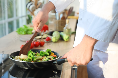 Male chef with spatula frying tasty vegetables in kitchen, closeup