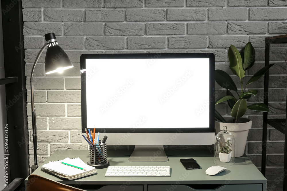 Fototapeta premium Workplace with computer, mobile phone, stationery supplies and glowing lamp near grey brick wall