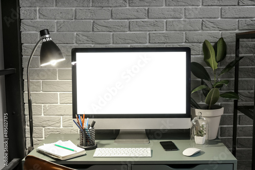 Workplace with computer, mobile phone, stationery supplies and glowing lamp near grey brick wall