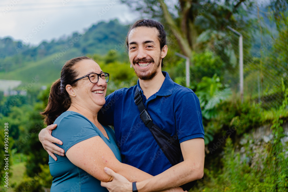 latin mother hugging her son young man with beard, outside in the forest. smiling with love.