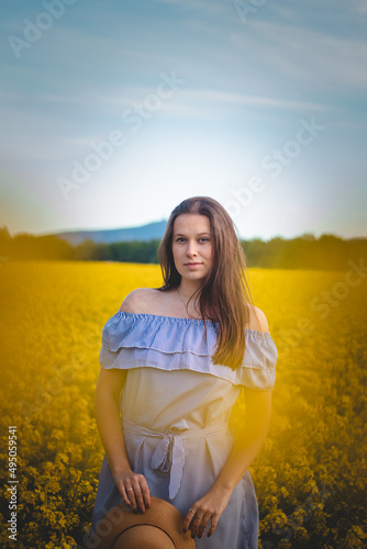 Breathtaking candid portrait of a young brunette in a beautiful summer blue dress at sunset in a rape field. The brown-haired model is smiling naturally. Fashion style