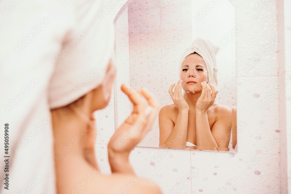 Young beautiful woman using a moisturizing facial mask after taking a bath. Pretty attractive girl in a towel on her head stands in front of a mirror in a home bathroom. Daily hygiene and skin care Young beautiful woman using a moisturizing facial mask after taking a bath. Pretty attractive girl in a towel on her head stands in front of a mirror in a home bathroom. Daily hygiene and skin care