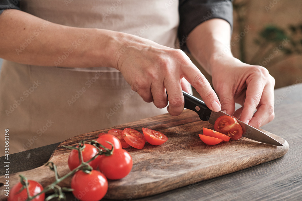 Close-up of female hands slicing cherry tomato with knife on wooden board, cooking vegetable salad Close-up of female hands slicing cherry tomato with knife on wooden board, cooking vegetable salad