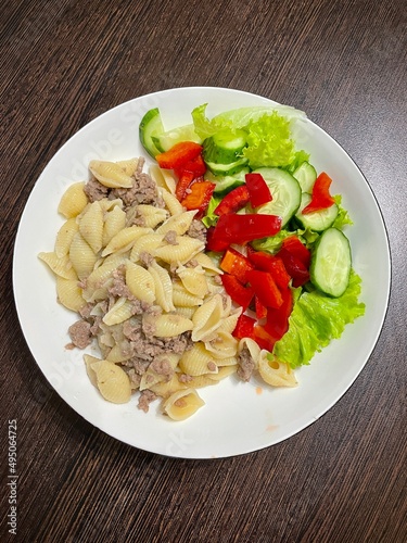 large vertical photo. homemade food, healthy useful products. round white plate on a brown wooden table. pasta with ground beef and cucumber, red bell pepper and cherry tomato salad. eco.